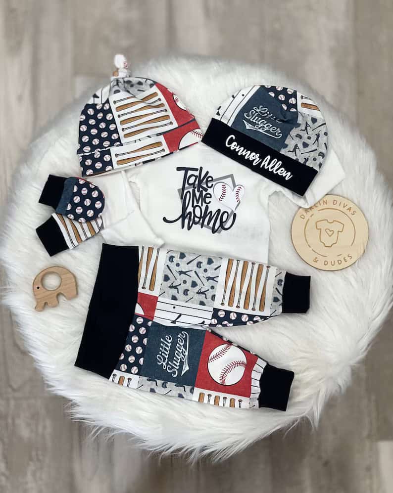 Infant outfit set on a round white rug. Perfect coming home outfits include a white onesie with Take Me Home, pants with baseball-themed patchwork, and matching hat. Accompanied by a wooden elephant toy and a round wooden sign with heart and bear illustration.