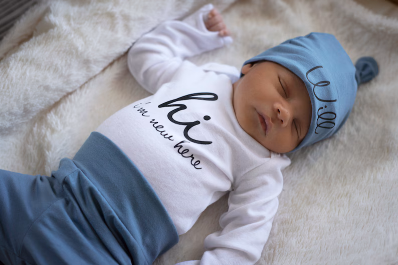 A newborn baby sleeps peacefully on a soft, white blanket, dressed in a charming coming home outfit with a blue hat and matching pants. The babys white shirt reads hi, Im new here across the front.