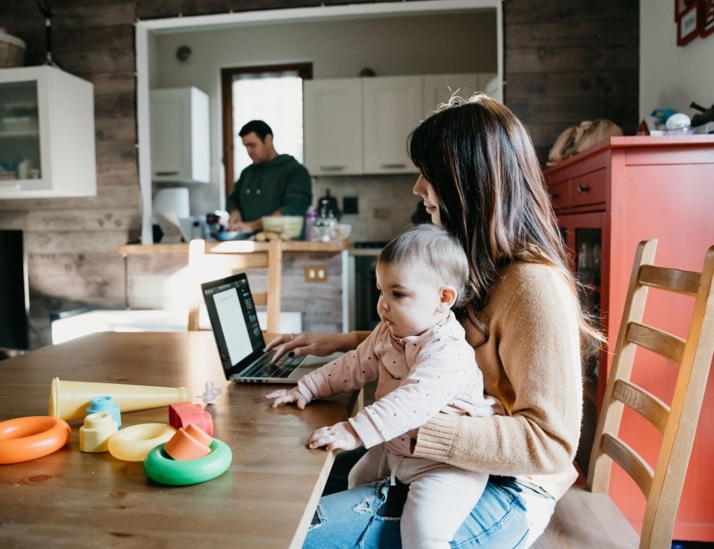 A little girl using a laptop with her mom while the dad is cooking in the kitchen. They are sitting at the table, the mother is holding her daughter.