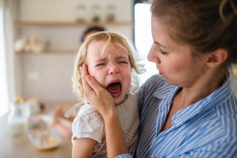 A mother holding a crying toddler daughter indoors in kitchen when cooking.