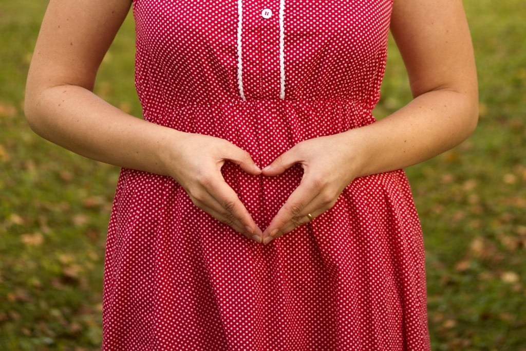 Woman forming a heart over her belly as a sign that she is pregnant but it is still a secret for the rest of the world. She is wearing a red dress with white dots and a ring on her finger.
