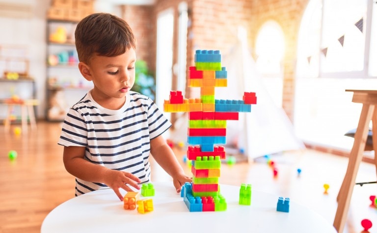 A young child wearing a striped shirt is standing at a round white table, focused on building a colorful structure using interlocking plastic bricks. The background shows a bright room with toys and a play tent, illustrating the child's ability to play independently.