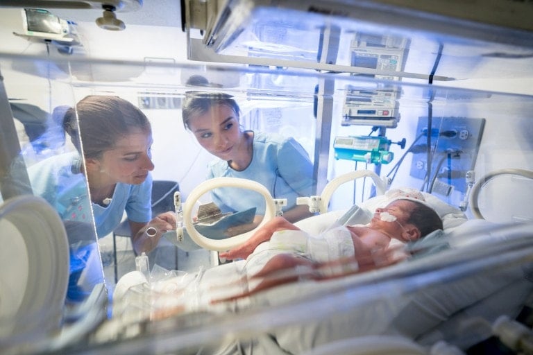 Concerned team of nurses in the intensive neonatal care unit looking at a premature newborn in an incubator while holding his medical chart on a clipboard.