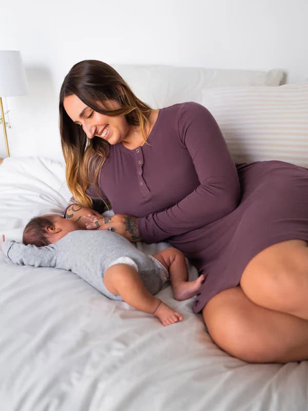 A woman in a mauve dress sits on a bed, breastfeeding her baby, dressed in gray. Smiling, she gazes lovingly at the infant. White and striped pillows fill the backdrop—a serene scene capturing intimate moments that inspire thoughts of perfect gifts to bring a new family.