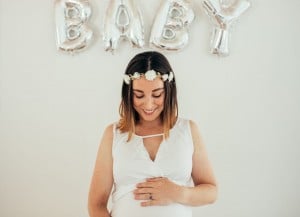 Expecting mother showing her pregnant belly while standing under a baby sign of balloons at her baby shower.