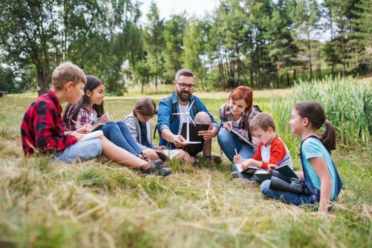 A group of children and two adults are sitting in a grassy area near trees, engaged in learning activities. The man is holding a tablet, and the children are writing or drawing in notebooks. The scene appears to be an outdoor educational session, showcasing alternatives to public school.