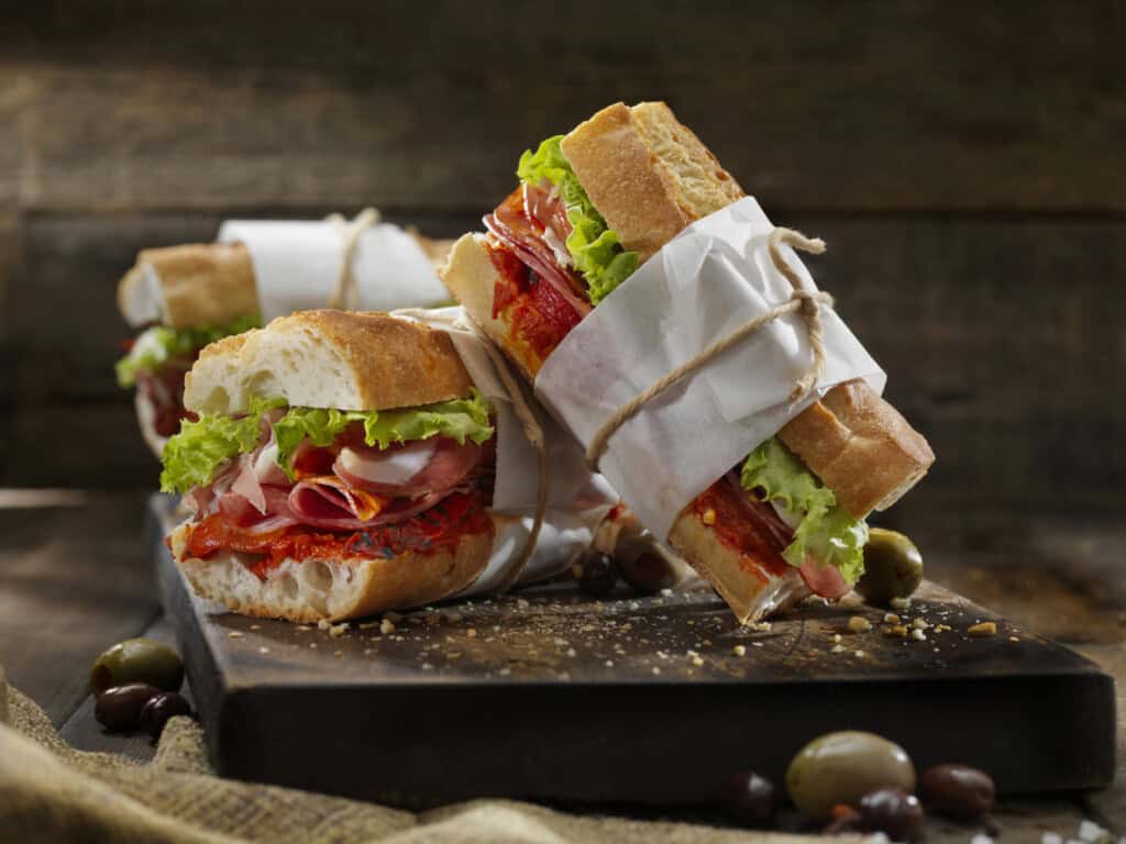 Three sandwiches with crusty bread, lettuce, and cured meats are wrapped in white paper and tied with twine like thoughtful gifts to bring a new family. They rest on a dark wooden surface, surrounded by scattered olives, set against a rustic wooden background.