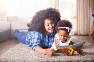An adult and a young child are lying on a carpeted floor, demonstrating how to teach a child to read. The adult, wearing a blue plaid shirt, smiles while the child, dressed in a white polka-dot dress with a bow, is intently looking at the book. A couch is visible in the background.