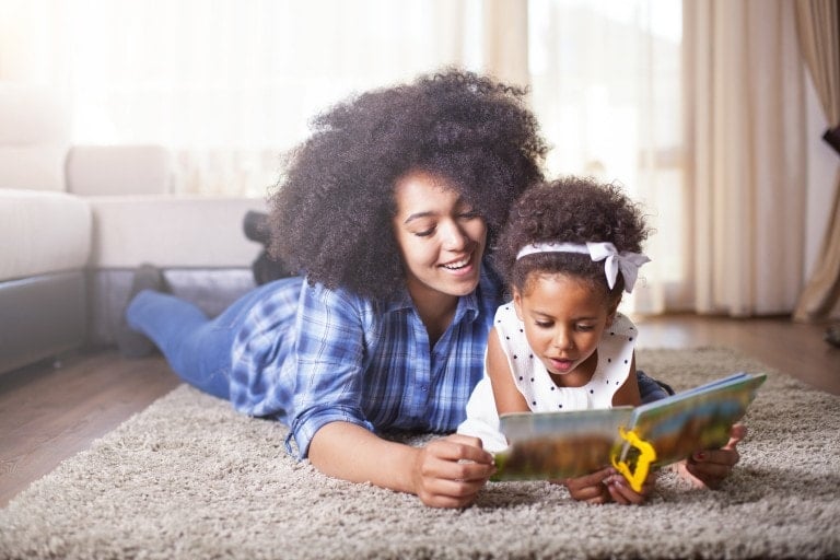 Mother reading a book to her daughter on carpet.