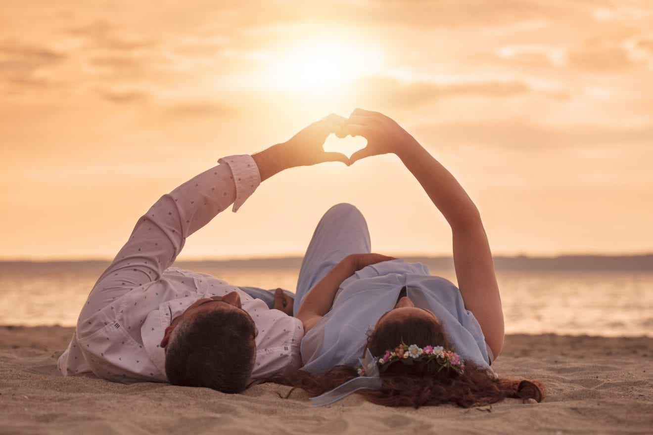 Pregnant couple lying on beach and making heart shape at sunset