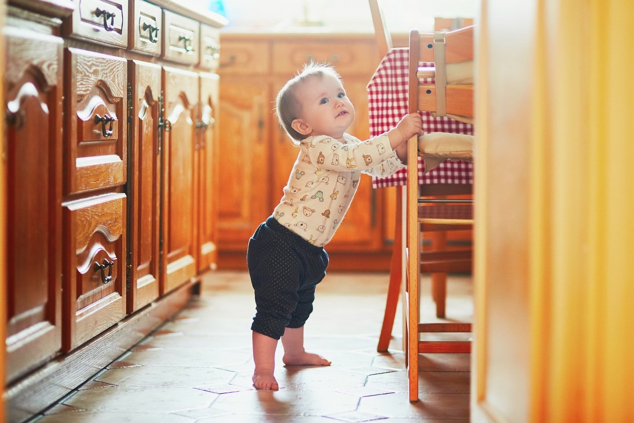 Baby girl standing on the floor in the kitchen and holding on to furniture. Little child pulling up at home.