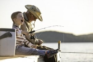 A grandfather is teaching his grandson to fish during sunset in summer. They are both sitting on the dock and laughing. It is a beautiful summer day. Across the lake, there is a mountain.
