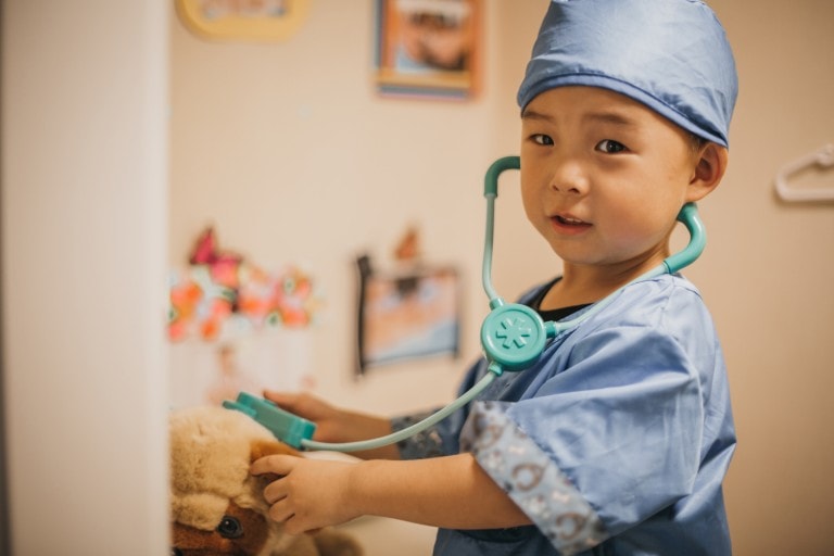 A young child dressed in blue scrubs and a blue cap is holding a stethoscope to a stuffed animal, engaged in an imaginative dress-up play session as a doctor. The room features colorful pictures on the wall in the background, enhancing the playful atmosphere.