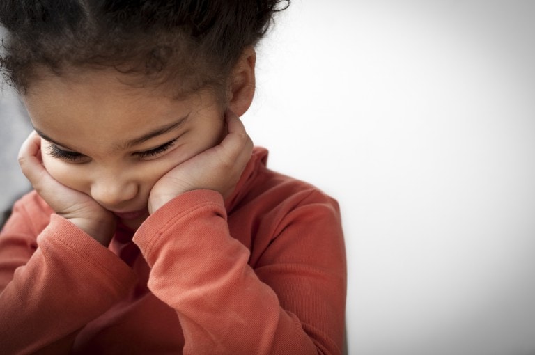 A young child with curly hair wearing an orange long-sleeve shirt is resting their head on their hands, appearing to be in deep thought or sadness, possibly experiencing child regression. The background is a plain, neutral color. The child's eyes are looking downward.