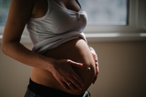 A pregnant person stands indoors wearing a white tank top lifted to reveal a round belly. Gently holding their belly with both hands, they seem to be exploring early labor positions. The background is softly lit by natural light from a window.