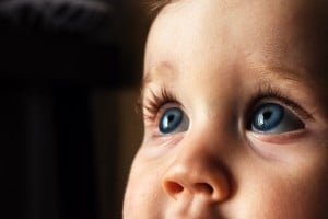 Close-up photograph of a young child's face, focusing on their large blue eyes and long eyelashes. The child is looking upwards with an expression of wonder, capturing the magic as babies' eyes change color over time. The background is dimly lit and out of focus.