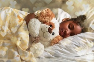 A child with brown hair in a bun is sleeping in a white bed, cuddling a brown and white stuffed dog. The child is covered with a yellow and white floral-patterned blanket. The background has a warm, heart-shaped bokeh effect, suggesting an early bedtime filled with love and comfort.