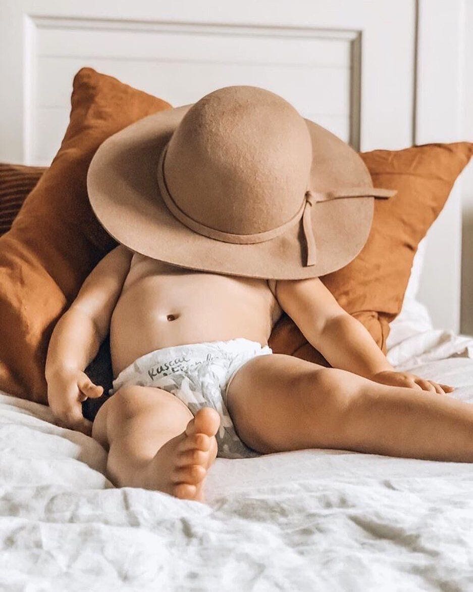 Baby laying down with a hat on its head wearing a training pant.