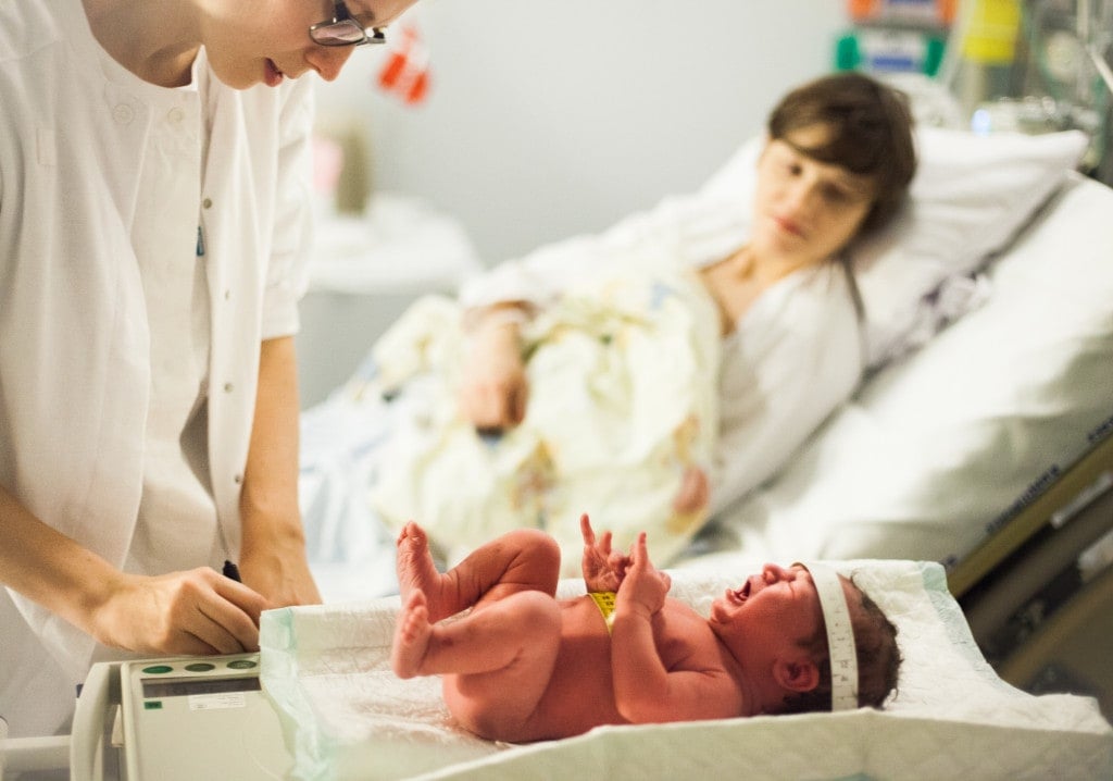 Calm and patient midwife weighs a screaming and crying newborn baby, mother in background