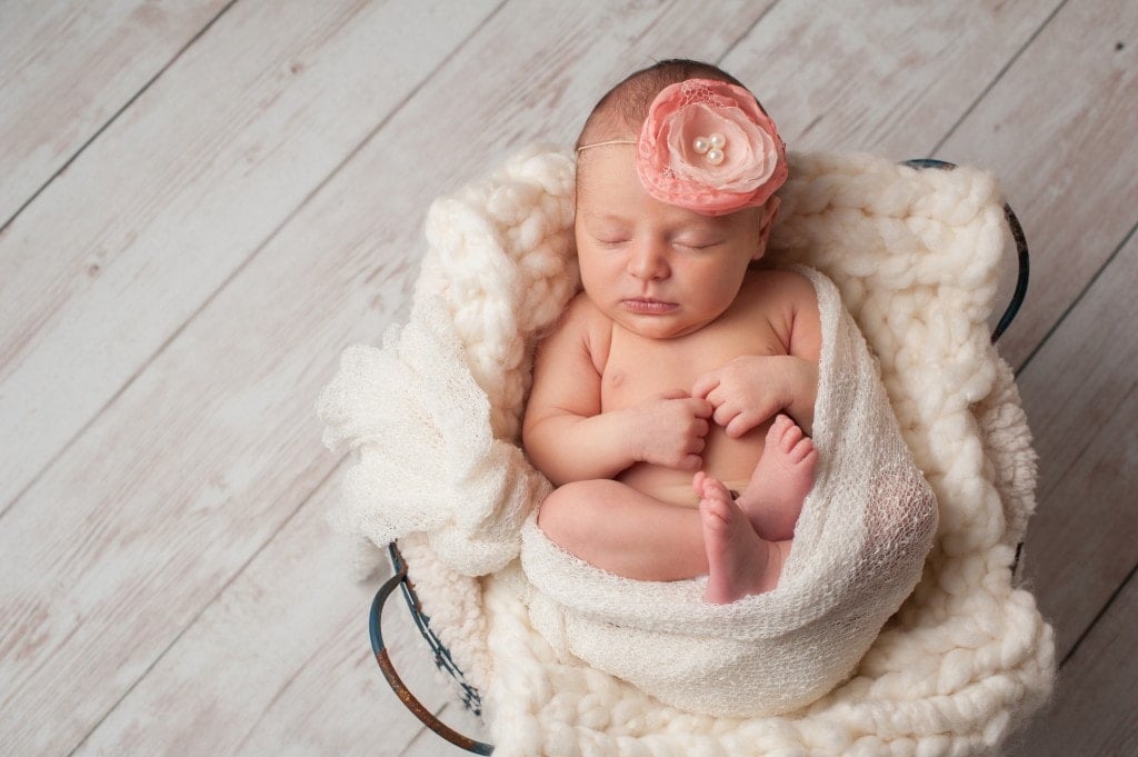 A portrait of a beautiful, seven day old, newborn baby girl wearing a large, fabric rose headband. She is swaddled with gauzy fabric and sleeping in a wire basket.