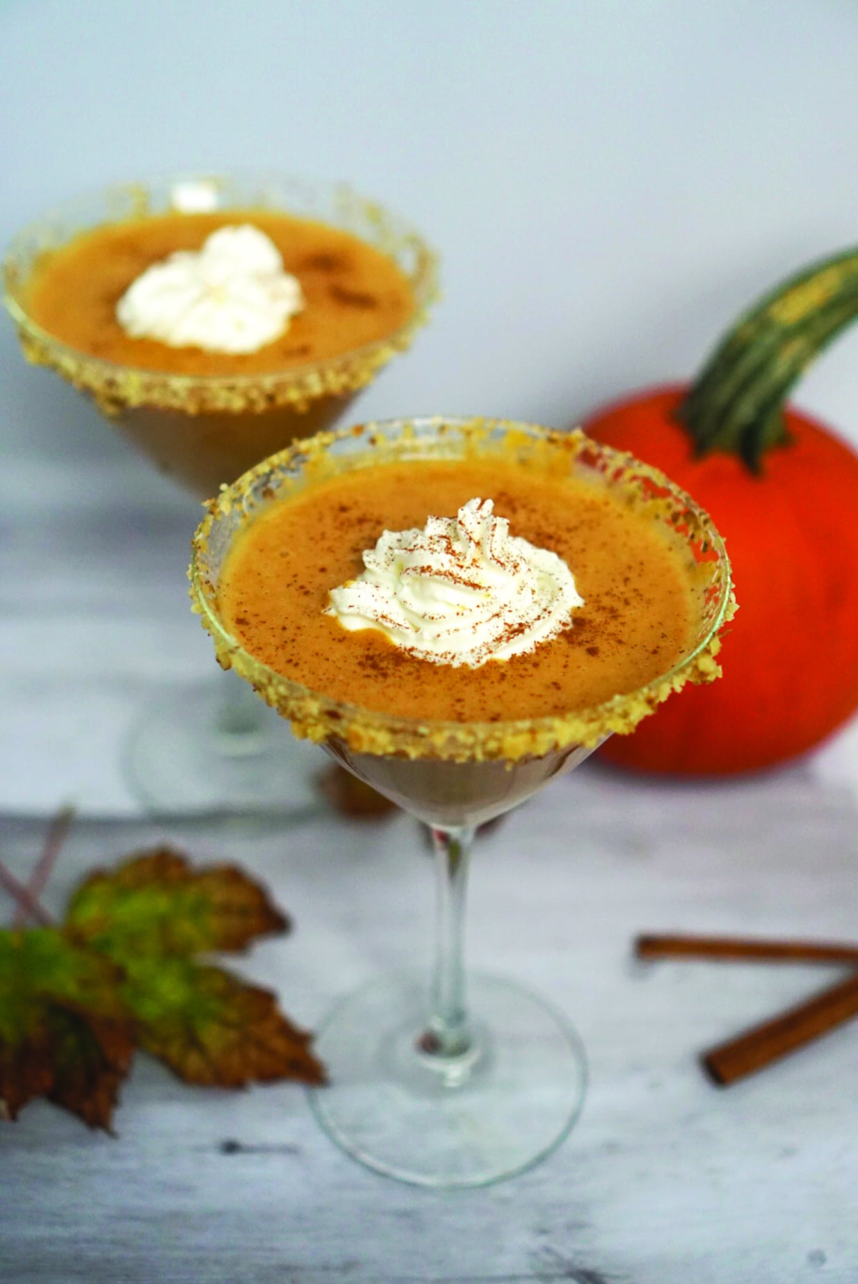 Two martini glasses filled with a creamy pumpkin-colored drink are garnished with whipped cream and sprinkled with spices. The glass rims are sugared, making them perfect for fall mocktail recipes. A whole pumpkin, dried leaves, and cinnamon sticks are visible in the background.