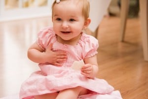 A baby wearing a pink dress sits on the floor, holding a cookie in one hand and touching her chest with the other, with a slight smile on her face.