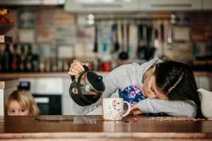 A woman, clearly exhausted, is sleeping with her head resting on a kitchen table while still pouring coffee from a French press into a mug. A small child is peeking over the edge of the table in the background. The kitchen has various utensils and a colorful backsplash.