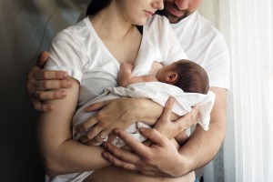 A couple standing close together, each wearing white shirts, while holding a newborn baby wrapped in a blanket. The woman cradles the baby, with the man's arms around both of them, symbolizing how dads help with breastfeeding. Natural light streams through the window, illuminating their tender moment.