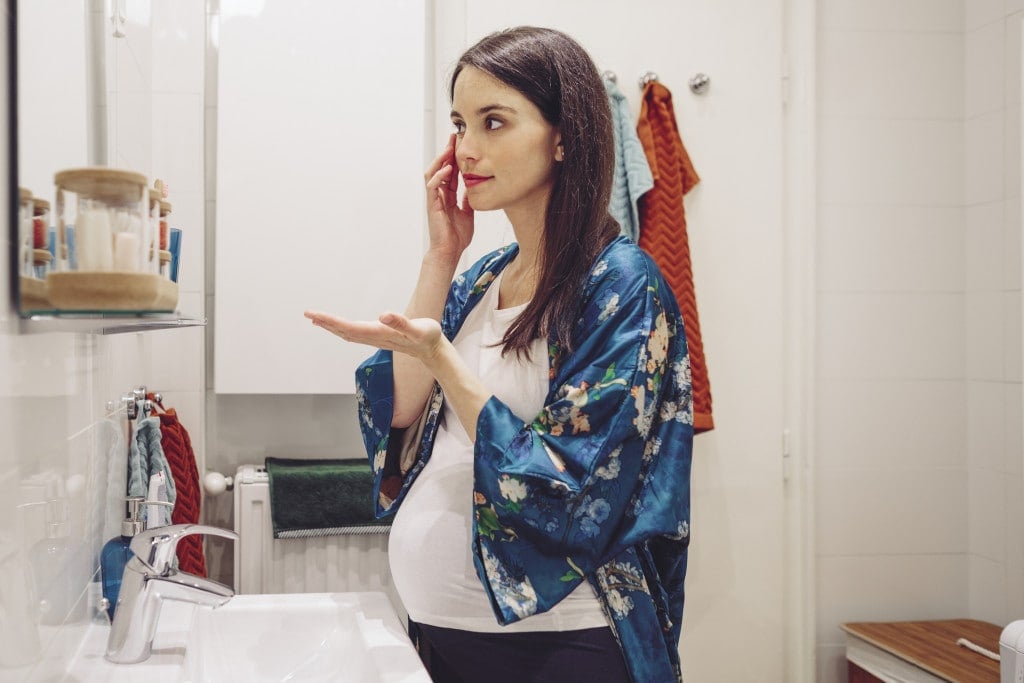 Pregnant woman standing in her bathroom doing some skin care to take care of melasma.