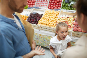 A girl with a surprised expression stands between two adults having a discussion at a fruit market. The stall displays fruits like apples, plums, and cucumbers, though a few look slightly spoiled. One of the adults holds a brown paper bag.