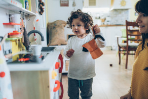 A young child plays with a toy kitchen set, holding a toy kettle while an adult watches and smiles in a home setting.