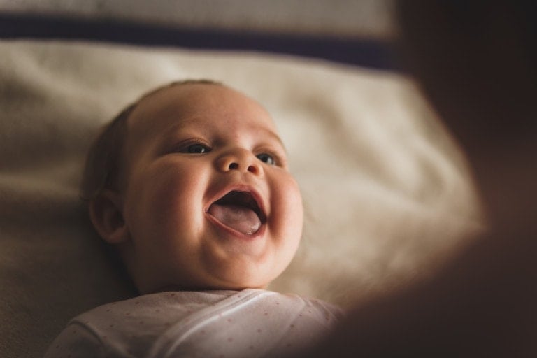 High angle view of cheerful newborn baby girl lying down on the bed and laughing while her mother, that is blurred in foreground, is playing with her.