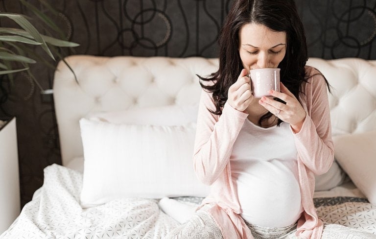 Beautiful young pregnant woman sitting in bedroom and drinking a cup of red raspberry leaf tea.