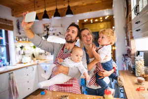 Beautiful young family making cookies at home. Father, mother, toddler boy and baby taking selfie with a smartphone.