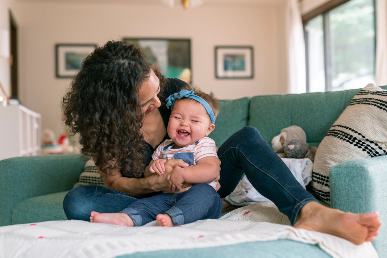 A woman with curly hair sits on a teal couch, holding a laughing baby in a blue headband and overalls. They are in a well-lit living room with framed pictures and large windows. A stuffed animal is beside them as if waiting to join the fun. Wondering how to make a baby laugh? This moment says it all.