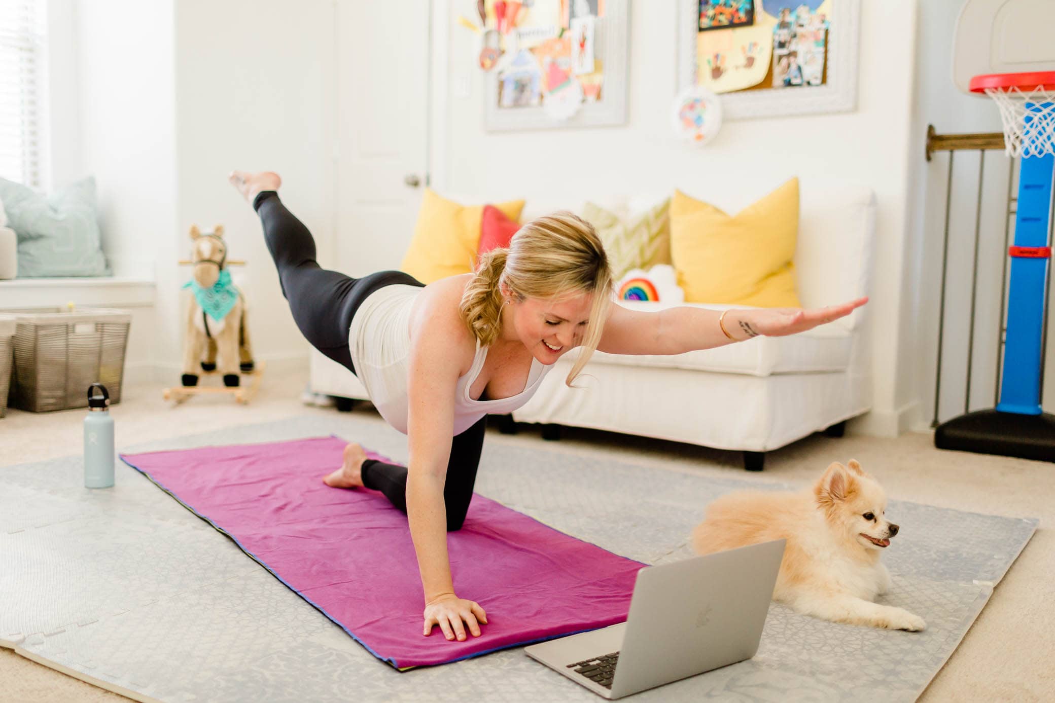 Pregnant woman on her yoga mat looking at her laptop doing a workout in her kids' playroom.