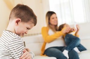 A young boy with a striped shirt stands in the foreground, looking upset with his arms crossed. In the background, an adult woman in a yellow sweater is sitting on a couch, holding and playing with another young child, capturing a moment of sibling jealousy.