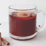 A glass mug filled with a dark liquid, possibly tea or hot cider, sits on a white surface. Inside the liquid, there are visible floating slices of fruit and spices. Cinnamon sticks are placed nearby on the table, evoking the cozy feel of a holiday mocktail.
