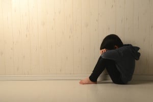 A young person sits on the floor in a corner, hugging their knees and resting their head on their arms. They are wearing a dark hoodie and pants, and the room has light-colored wooden walls and floor. The person appears to be alone, trying to cope with change in a contemplative posture.