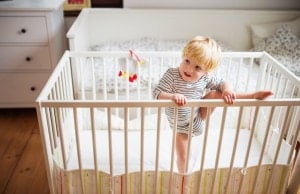 A toddler in a striped onesie is attempting to climb over the white bars of a crib. The crib is situated in a well-lit room with white furniture, including a chest of drawers on the left and a big kid bed with patterned bedding in the background.
