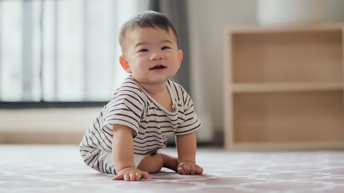 A baby wearing a striped onesie is crawling on a light-colored carpet in a room with a window and a wooden shelf in the background. With short dark hair and an adorable smile, its almost as if the baby has just experienced one of those precious moments when do babies laugh.