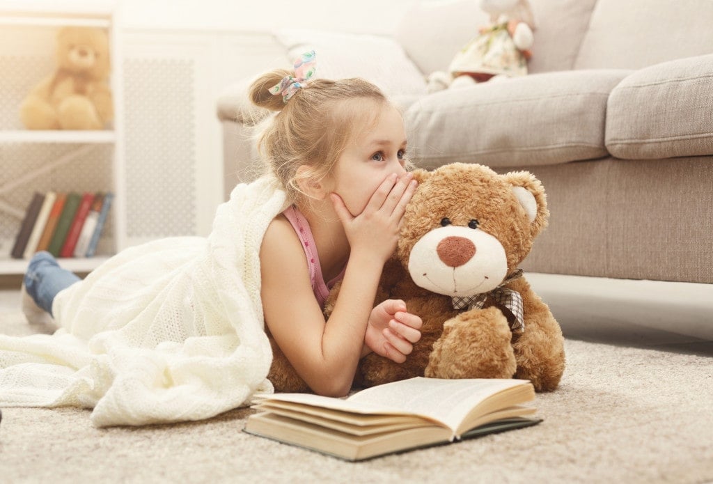 Cute happy little casual girl embracing teddy bear, reading book and sharing secrets with her favorite toy friend.