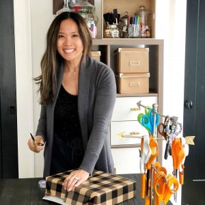 A person is standing at a table and wrapping a gift with checkered wrapping paper. They are holding a pair of scissors and smiling at the camera. Behind them is a shelf with various craft supplies and storage boxes. A container with scissors is on the table.