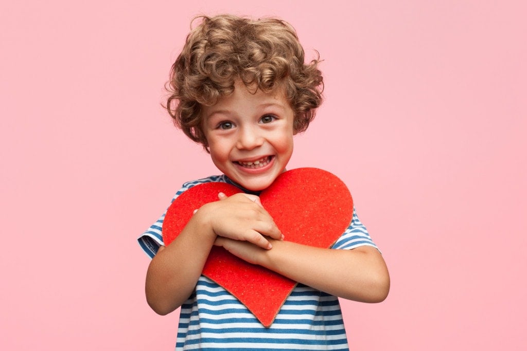 Little curly boy holding heart application and laughing at camera on pink background.