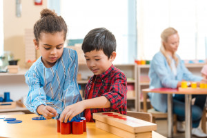 Two children work together with geometric shapes at a classroom table, while two adults interact in the background.