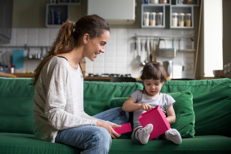 A woman and a young child sit on a green couch. The woman is holding an open pink box, which appears to be filled with items not to buy for toddlers, and the child is examining its contents. The background shows a kitchen with white tiles, various utensils, and jars on shelves.