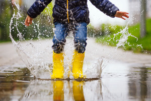 A child wearing yellow rain boots and a dark jacket jumps into a large puddle on a wet sidewalk, causing water to splash up around them. The blurred background emphasizes the child's motion, capturing the joy of a rainy day.