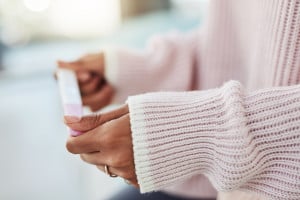 A person wearing a pink knitted sweater is holding a pregnancy test with both hands, perhaps taking it too early. The scene is indoors with a blurred background, focusing on the person's hands and the test.