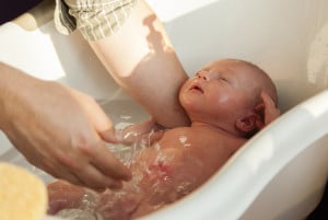 A person is giving a babies bath in a white baby bathtub. The newborn lies on its back with eyes closed, one arm resting on the side of the tub. The adult's arm supports the baby and pours water over them. A sponge is visible on the left side.