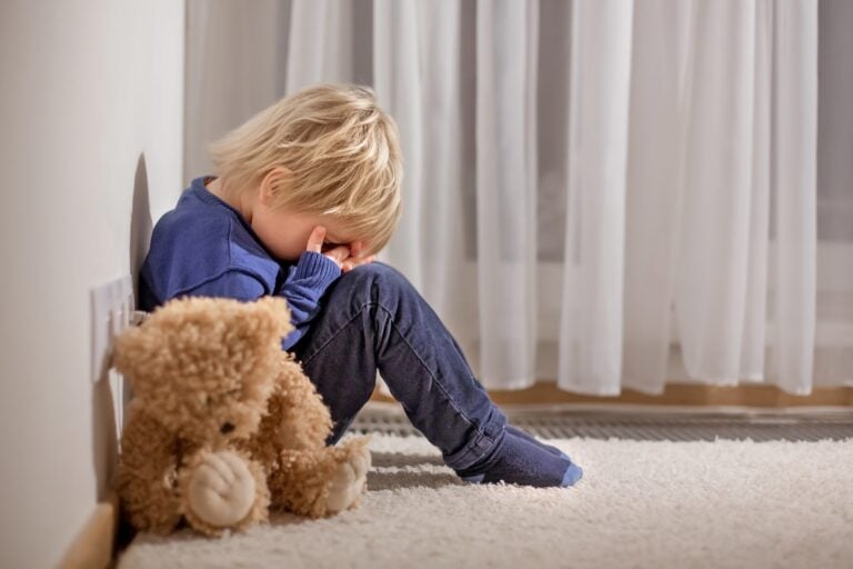A child with blond hair sits on the floor against a wall, covering their face with their hands in a moment of quiet introspection. The child is wearing a blue long-sleeve shirt and dark pants, likely seeking comfort. A brown teddy bear lies beside them, while white curtains softly frame the background.